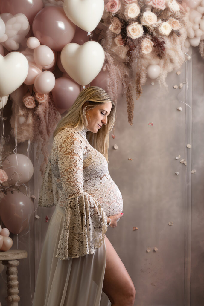 Zwangere vrouw in zand kleurig kanten jurk voor een achtergrond van zacht roze hart vorige ballonnen tijdens een zwangerschap fotoshoot bij Little Miracle memories