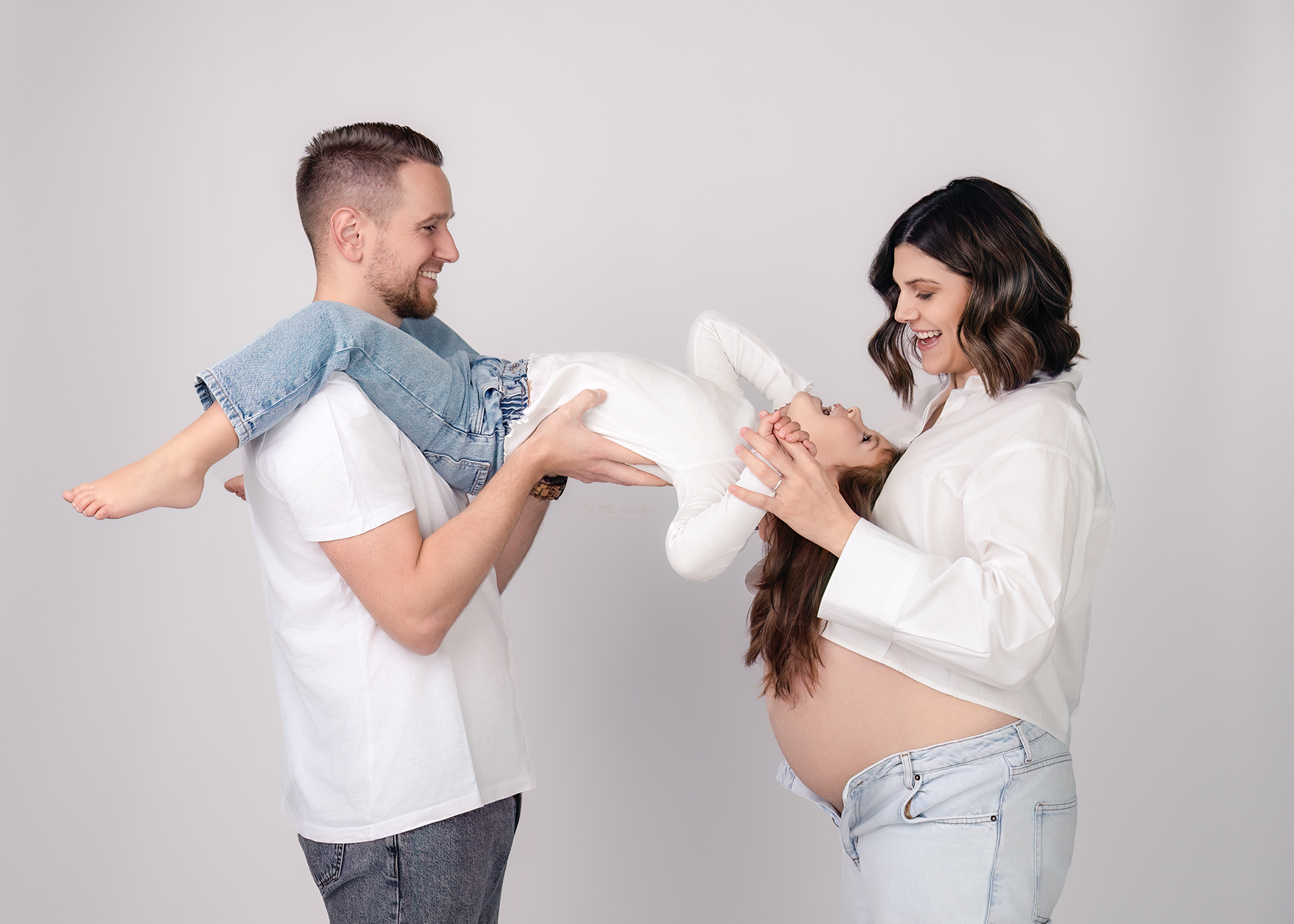 Zwangerschapsfoto van een gezin met mama, papa en kind, liefdevol verbonden rond de zwangere buik, gefotografeerd in een lichte studio in Zandhoven